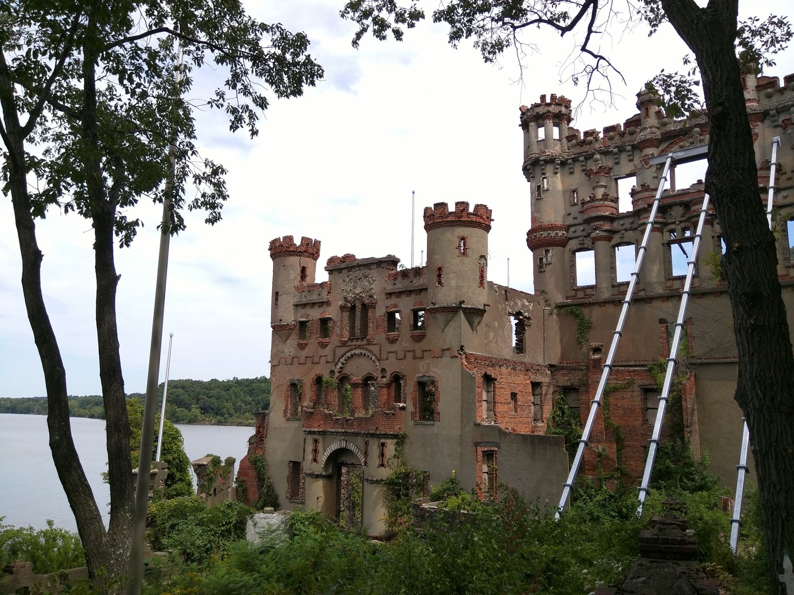 A ruined castle with scaffolding stands near a body of water, framed by trees.
