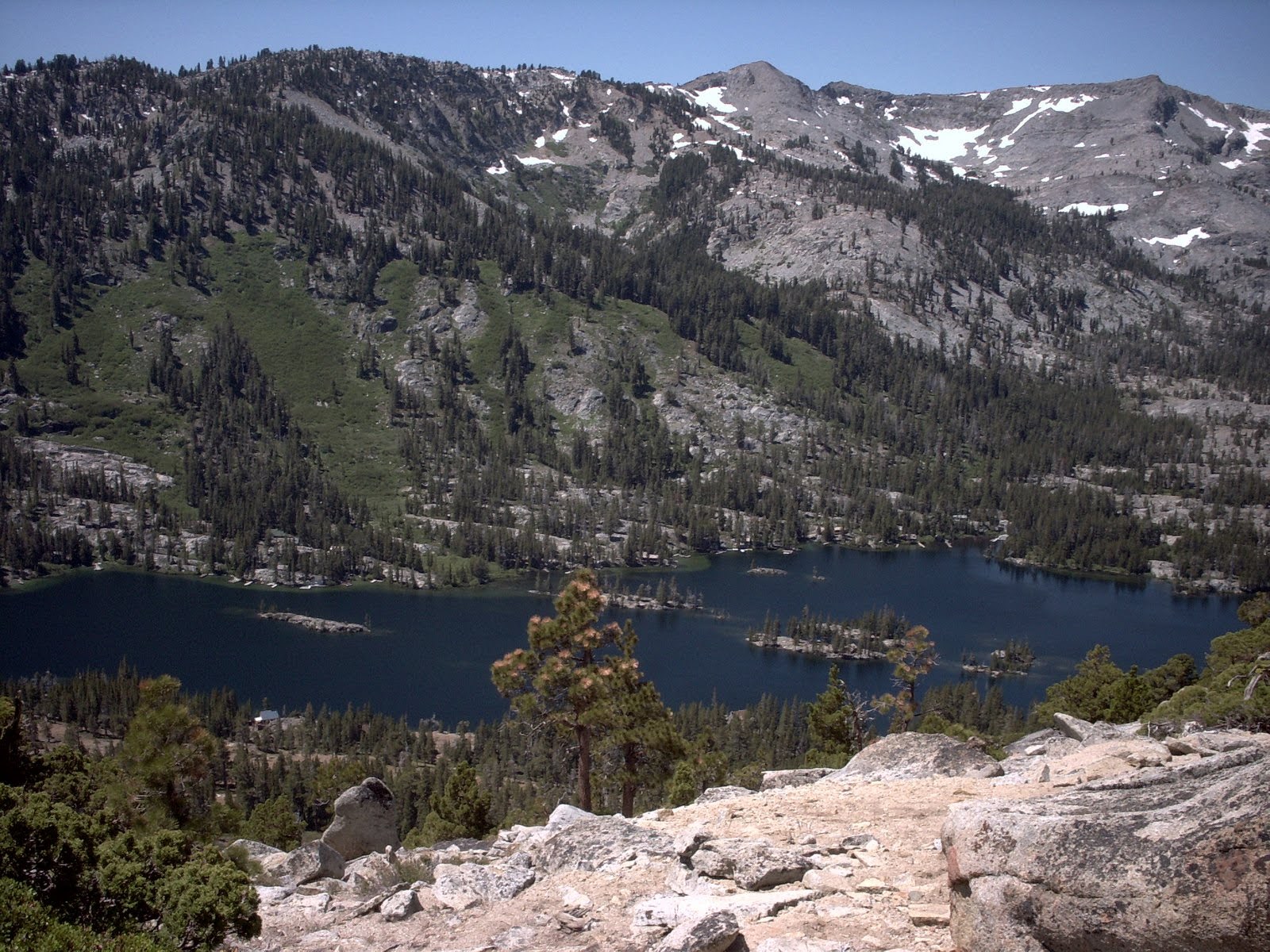 A high-altitude mountain lake surrounded by coniferous forests and rocky slopes with patches of snow.