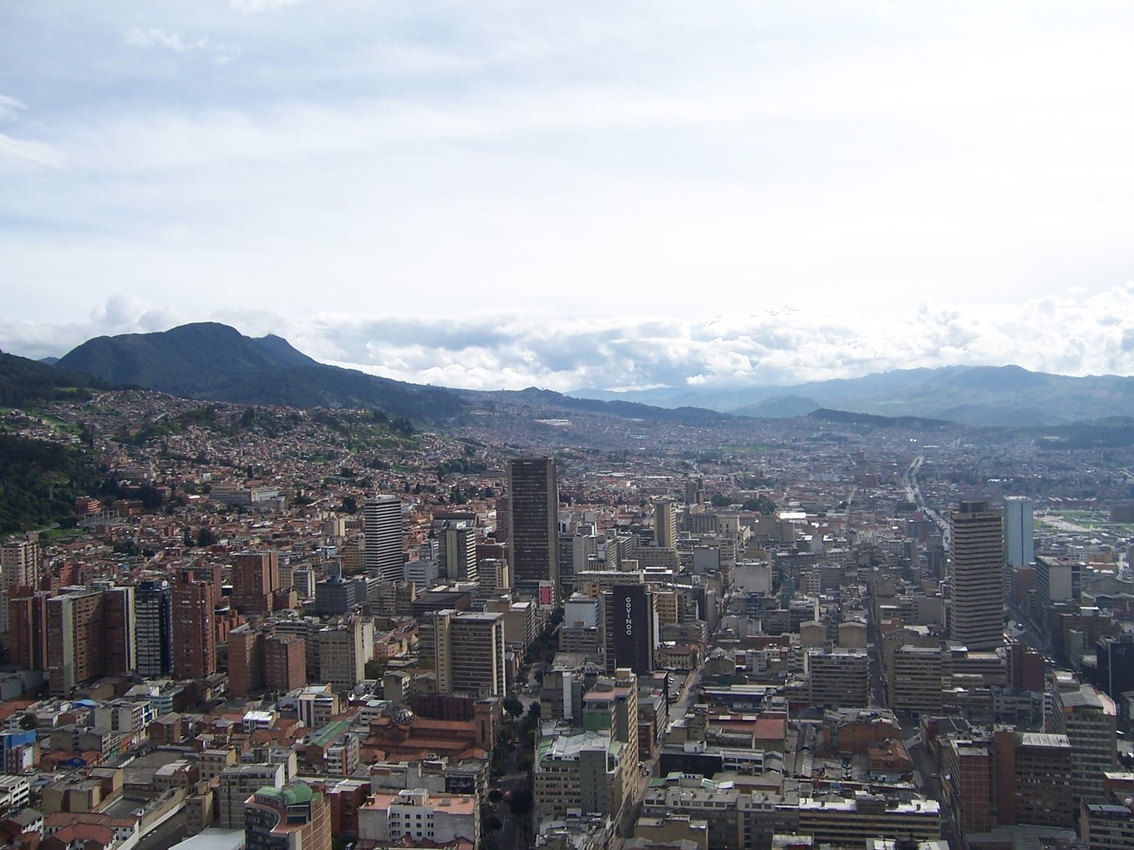 A high-angle view of a dense city with high-rise buildings nestled against a mountain range under a cloudy sky.